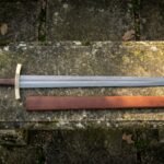 Hand forged Damascus medieval sword with a rosewood grip, bone cross and faceted brass pommel, lying with its brown leather scabbard on a mossy stone bench.