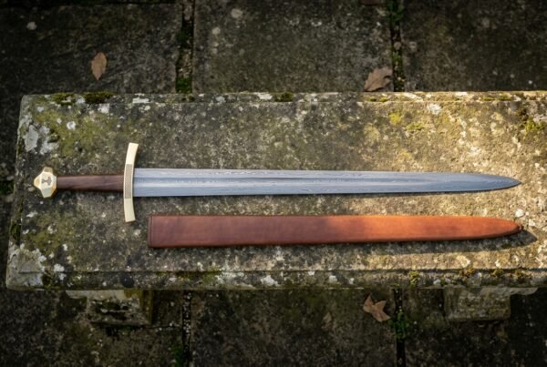 Hand forged Damascus medieval sword with a rosewood grip, bone cross and faceted brass pommel, lying with its brown leather scabbard on a mossy stone bench.