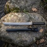 Hand forged tanto with a black cord wrapped grip and square iron tsuba resting on a lichen covered boulder, its black lacquered saya lying below the blade