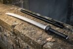 Hand forged katana with black cord grip and blackened iron tsuba resting on a stone ledge against a dark timber door, black saya lying behind the blade