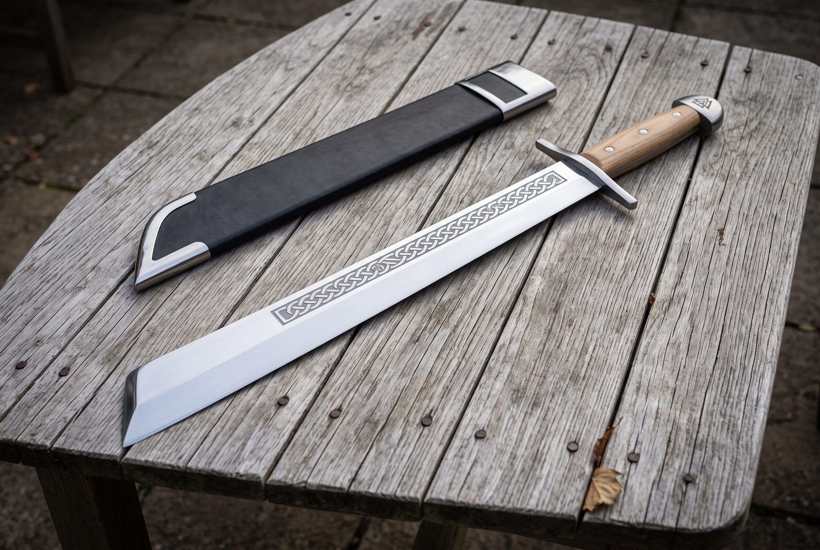 Gemini_Generated_Image_uo6h5uo6h5uo6h5u Hand forged grosse messer sword with a broad polished single edged blade and pale hardwood grip lying on a silvered timber table beside a black leather sheath
