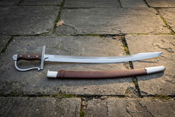 Hand forged pirate cutlass with a broad clipped point blade, fullered forte, steel knuckle bow and walnut slab grip laid on grey flagstone paving beside its brown leather scabbard