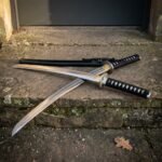 Battle ready hand forged katana crossed over its black lacquered saya on a weathered stone step, black cord grip and brass toned tsuba lit from the side