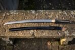 Hand forged katana with a black cord wrapped tsuka and black lacquered saya laid along a lichen covered stone bench in soft low winter light