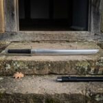 Hand forged dao sword with a straight backed polished single edged blade and black cord wrapped grip lying on a stone doorstep beside its black scabbard