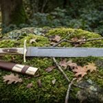 Hand forged Viking seax with a layered Damascus blade and stag antler handle resting on a moss covered boulder in autumn woodland, brown leather sheath beside it