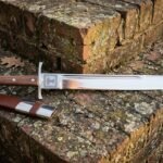 Hand forged grosse messer with a long polished straight backed blade and rosewood grip resting on a lichened stone ledge beside a brown leather sheath