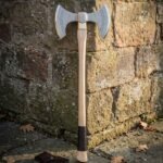 Hand forged double headed axe with two symmetric etched steel cutting edges and a light wood haft wrapped in dark leather, standing against a red brick wall