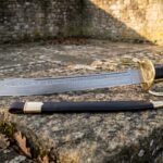 Hand forged Damascus naval cutlass with a layered rune engraved blade, ornate brass shell guard and black ribbed grip lying on a weathered stone ledge beside its black leather scabbard