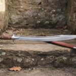 Hand forged tulwar with a satin finished curved blade, rosewood grip and steel disc pommel lying on a mossy stone step beside its russet leather scabbard