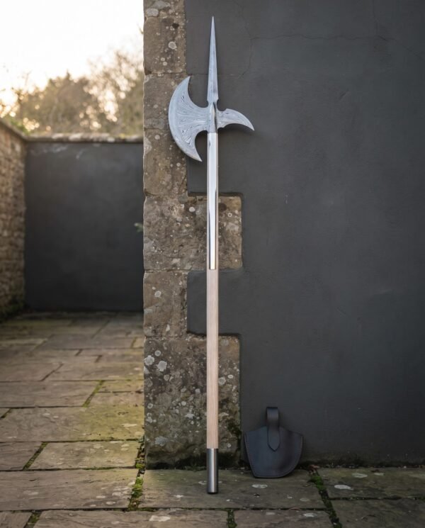 Damascus halberd with a forge welded spike, patterned crescent blade and hooked fluke on a pale ash haft standing against a stone and slate garden wall