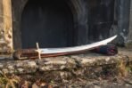 Ancient Persian sword pattern hand forged as a shamshir with a deeply curved polished blade and carved vine scroll grip on a stone ledge before an arched doorway