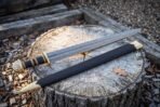 Angled view of a Damascus Viking sword with brass guard and brass banded grip lying with its scabbard on a stump