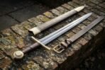 Arming sword with a Damascus blade, brass disc pommel and brown leather grip beside its brass tipped runed scabbard and buckled belt on brick