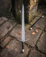 Arming sword with a Damascus blade and quatrefoil tipped guard propped against a stone wall, black leather scabbard with brass throat below