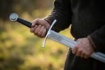 Arming sword hilt with curved crossguard, wheel pommel and black grip held in both hands above the knotwork etched blade