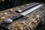 Arming sword with black leather grip, straight cross guard and wheel pommel laid on a mossy stone bench beside its black scabbard