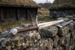 Arming sword with brown leather grip and engraved disc pommel on a dry stone wall beside its black leather scabbard