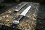 Arming sword with engraved wheel pommel and banded black leather grip resting on a stone ledge with its scabbard behind