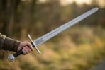 Arming sword with a fullered steel blade, knotwork panel and black grip held out at arm's length over a sunlit field