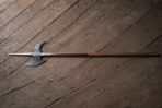 Battle halberd with a satin steel spike, crescent blade and hooked fluke on a long hardwood haft laid across a dark weathered plank floor