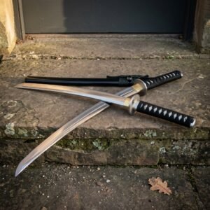 Battle ready hand forged katana crossed over its black lacquered saya on a weathered stone step, black cord grip and brass toned tsuba lit from the side