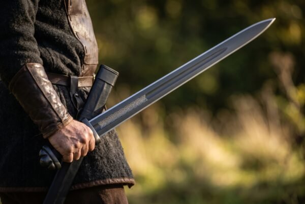 Black Damascus Viking sword with a dark fullered blade held point up beside its black scabbard by a man in a leather bracer