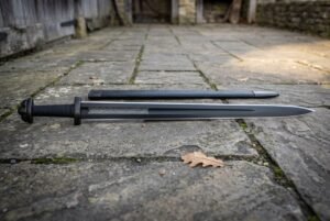 Black Damascus Viking sword with dark etched layered blade and an inscription across the face, resting on grey flagstone paving beside its black scabbard