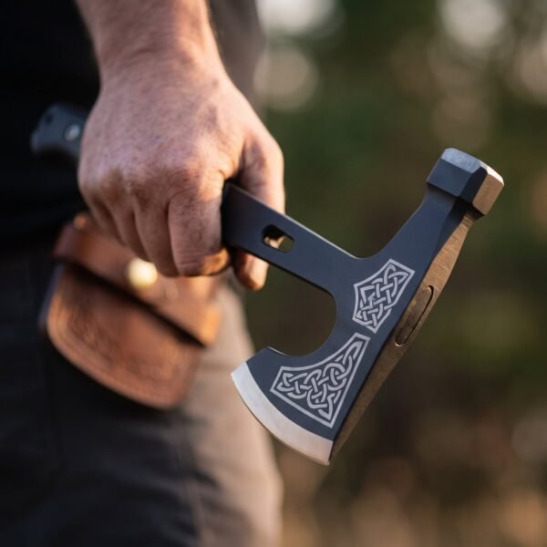 Black tactical tomahawk with Celtic knotwork etched head and polished edge held in a hand beside a tooled leather sheath
