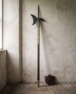 Blackened halberd polearm with an axial spike and crescent blade on a walnut haft with brass langets standing against a pale cracked plaster wall