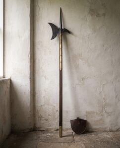Blackened halberd polearm with an axial spike and crescent blade on a walnut haft with brass langets standing against a pale cracked plaster wall