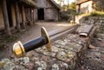 Brass pommel and guard of a Damascus falchion with black grip in close focus on an old brick wall, scabbard behind