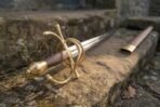 Brass swept hilt dueling sword with wood grip resting on a mossy stone step beside its leather scabbard