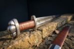 Close up of a broadsword hilt with a lobed steel pommel, knurled guard and russet leather grip, blade runes fading into the background