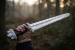 Broadsword with a rune engraved high carbon steel blade held up in a gloved hand, lobed pommel and knurled guard in low sunlight