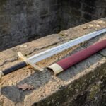 Camelot sword replica with an ornate gilt brass hilt, disc pommel and maroon leather scabbard resting on a weathered drystone wall in daylight