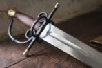 Close up of an espada ropera hilt with blackened side rings, brass tipped quillons and a wooden grip over a fullered steel blade