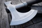 Close up of a bearded axe head with engraved rune bands, knotwork steel collar and dark wooden haft on a weathered table