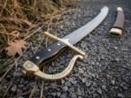 Close up of a brass knuckle bow cut with runes and a black horn grip on a Damascus sabre resting on gravel and dry grass