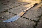Close up of a curved Damascus glaive blade with its steel socket and pale ash haft on a stone path