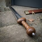Close up of a gladius hilt showing the ribbed wood grip, wooden guard block and globular pommel with steel peen on stone
