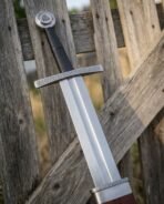 Close up of a longsword hilt with knotwork engraved steel cross, black leather grip and Valknut disc pommel against a fence