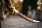 Close up of a shamshir hilt in a gloved hand above its basketweave tooled leather scabbard with brass chape