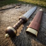Close up of the ribbed wooden grip, globular pommel and brass collars of a Damascus steel gladius on a tree stump by its scabbard