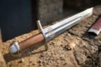 Close up of the steel cross guard, etched medallion and wooden grip of a high carbon steel falchion resting on warm sandstone