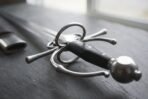 Close up of the swept ring hilt, pinned black grip and round steel pommel of a renaissance side sword on grey slate