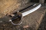 Close up of the three bar guard and ribbed black grip of a steel cavalry sabre with scabbard on stone