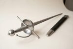 Close view of a polished cup hilt rapier with wire wrapped grip, round pommel and knuckle bow next to a black scabbard
