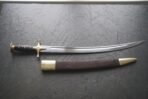 Curved high carbon steel talwar with black wooden grip, brass guard and disc pommel above its leather scabbard on slate