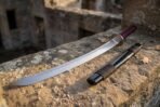 Curved polished steel blade of a short Japanese sword with red cord handle next to a black saya with brown cord bands